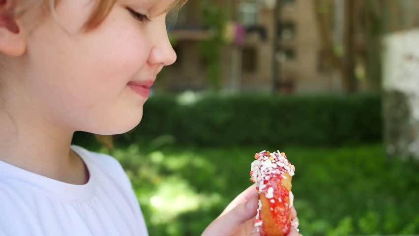 Portrait of a cute little teenage girl biting a sweet donut covered in jam and icing. Child eating donut outdoors fun and happy Royalty-Free Stock Footage #1104917785