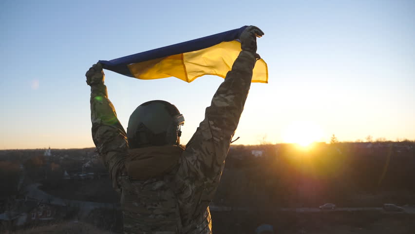Dolly shot of female soldier of ukrainian army a raised over head blue-yellow banner at sunset. Young girl in camouflage uniform lifted flag of Ukraine at countryside. Invasion resistance concept Royalty-Free Stock Footage #1104927749