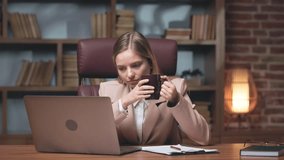 Female worker talking on video call on wireless personal computer and drinking cup of hot aromatic coffee. Exhausted woman feeling need in energy recovery during intensive working day.