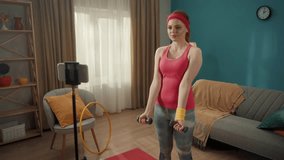 Young fit woman doing bicep exercises with dumbbells at home in front of a phone mounted on a tripod. The woman focused on lifting dumbbells to strengthen her arm muscles.