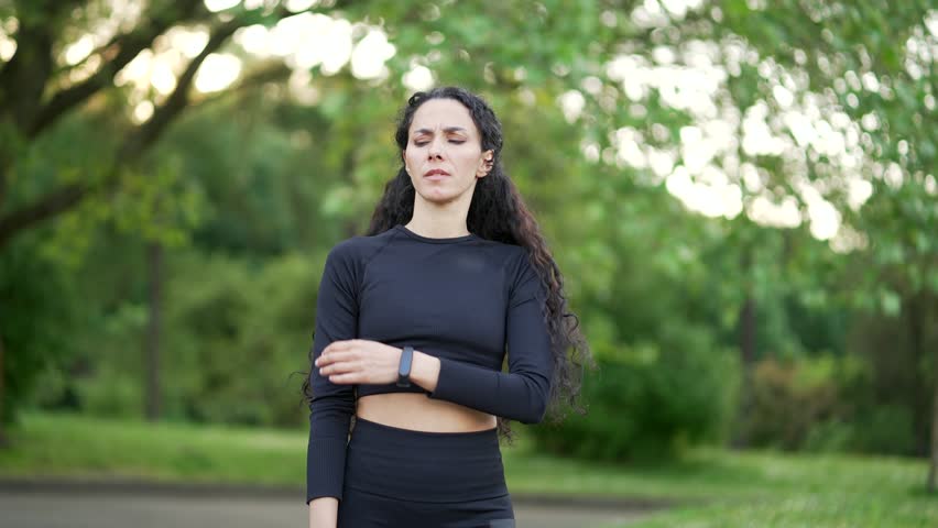 Young athletic woman in a tracksuit sharp pain in her shoulder during a morning jog in an urban city park. A sad female was injured. Unfortunate athlete rubs and massages the muscles on the sore spot Royalty-Free Stock Footage #1104949659