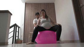 husband helps his pregnant wife to exercise on a ball at home