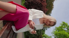 Vertical Video, Happy Attractive Blonde Woman in White Jacket and Fuchsia Skirt Sits on Bench in City Park and Uses Mobile Phone, Chatting with Friends on Internet and Social Networks. Slow Motion