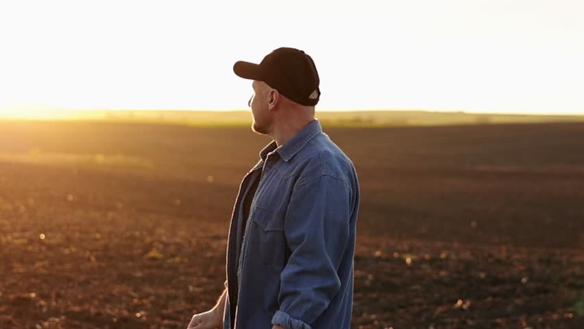 Side view of male farmer standing and looking in cultivated plowed field at sunset in spring. Owner agricultural farm is checking and examining farmland before sowing agriculture crops. Agribusiness Royalty-Free Stock Footage #1104951603