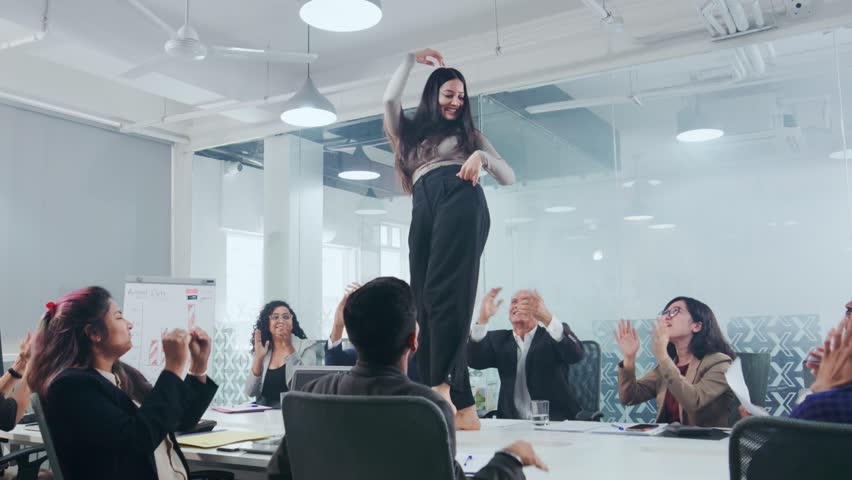 A happy Indian Asian modern corporate office executive woman employee enjoying and dancing while standing at the conference table while associates are busy or consumed in work in a board room meeting