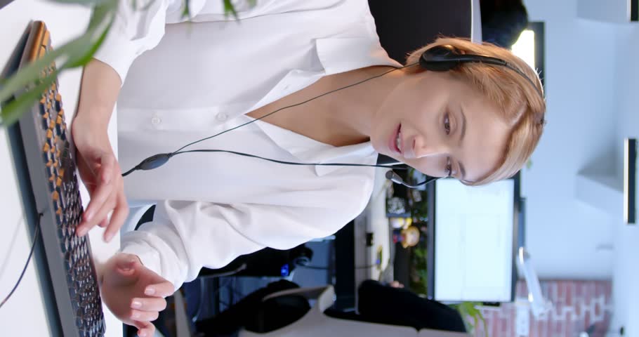Vertical shoot. Female manager at office. Woman at workplace is clapping keyboard and looking at computer. Middle-aged woman is smiling and communicate by headphones with microphone.