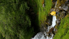 Gudvangen-Naeroyfjorden, Norway. Vertical Footage Video. Waterfall Tuftofossen In Spring. Young Caucasian Woman Lady Tourist Traveler Photographer Hands Up Enjoying Life.