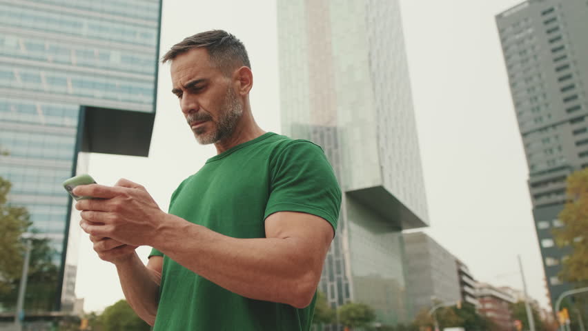 Mature man wearing casual clothes, standing on modern buildings background, texting on smartphone