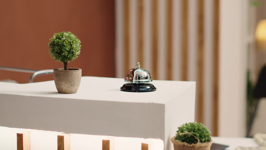 Elegant concierge bell on modern stylish hotel lobby check in desk. Reception bell next to mini plant on empty warm hospitality industry lounge reception counter, close up Royalty-Free Stock Footage #1104965533