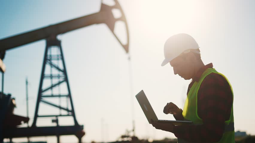 oil business. a worker works next to an oil pump holding a laptop. industry business oil and gas concept. lifestyle engineer studying the level of oil production on a laptop silhouette at sunset Royalty-Free Stock Footage #1104973821