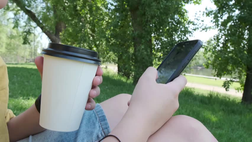 Mans hand type touchscreen of smartphone and held cup of coffee by another hand. Summer park background. 