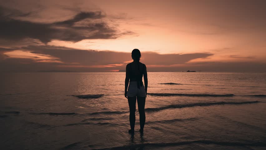 Silhouette woman looks at sunset, raises hands and enjoys beautiful sea sunset during vacation. Rear view happy woman standing on seashore, admiring beautiful sunset, enjoying relaxation and freedom
