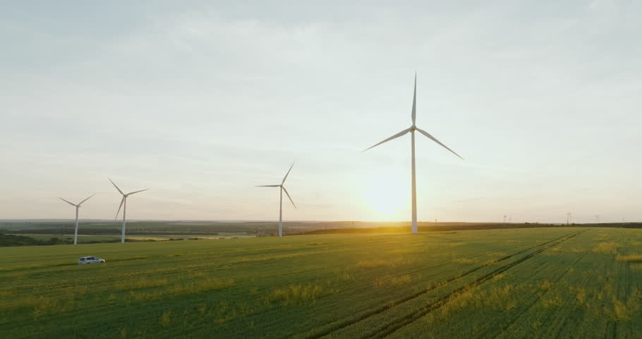 Large wind turbines against blue sky at sunset in spring. Auto car drives along rural road kicking up dust among green wheat field aerial view. Wind park agricultural farm. Alternative energy eco Royalty-Free Stock Footage #1104986715