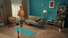 A man in sportswear is filming his home workout using a smartphone mounted on a tripod monopad. A man is jumping rope in the living room. Home fitness concept, online sports class. Slow motion.