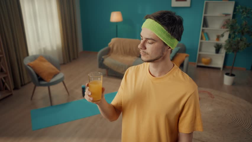 Portrait of a young man with a headband after a home workout. A man is happy to drink freshly squeezed juice, fresh against the backdrop of a living room with attributes for home fitness. Slow motion.