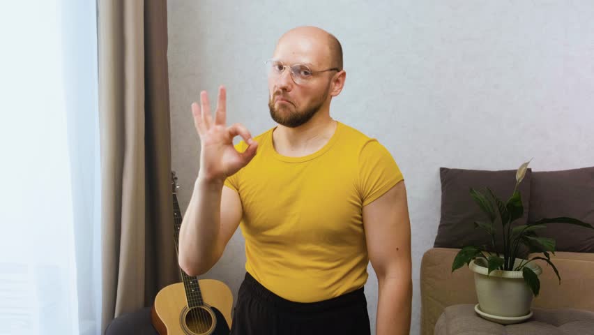 Funny bald and bearded nerd in glasses with high-stretched shorts muscles humorously nods approvingly and shows the OK gesture in the apartment at home