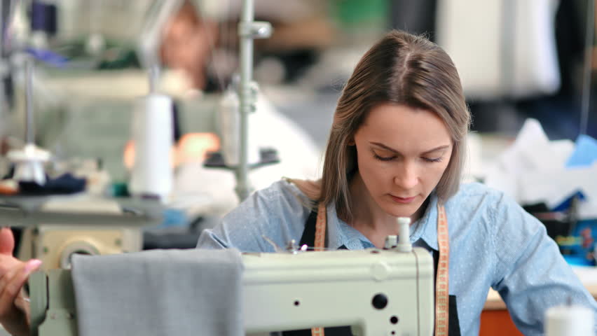 Focused woman seamstress professional dressmaker work use electric sewing machine at tailoring atelier. Female clothes designer occupation at industrial fabric garment manufacture needlework workshop