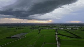 Video footage, aerial drone view, thunderstorms, dark clouds, low clouds, grassy fields, green rice fields, reservoirs, roads, before sunset, mountain passes, rural villages in Thailand.