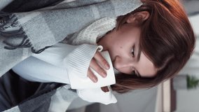 Vertical video. Throat drinking. Tea warm. Flu shivering. Cold brunette woman in blanket with hot beverage feeling frozen feeling pain sitting on couch in light home interior.