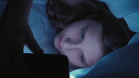 Vertical video. Screen time. Child insomnia. Late night. Internet addiction. Sleepless tired girl texting communicating on smartphone hiding under blanket in bed at home.