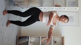 Vertical video. Squats exercise. Home workout. Fitness blog. Young active woman in sportswear training bending knees and reaching hands forward in light room interior.