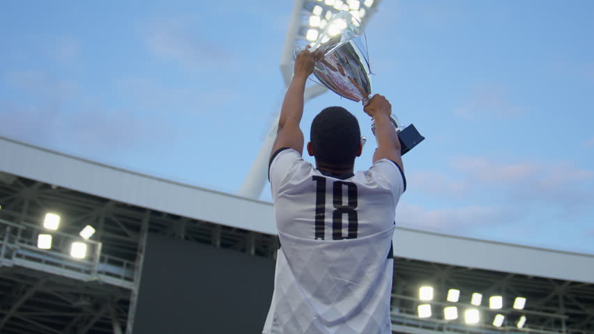 Portrait of African Black male soccer football player celebrating victory in the championship, lifting the trophy above his head in a huge stadium. Super slow motion shot