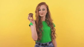 A beautiful young woman in a green t-shirt is standing in front of the camera and eating a cupcake. A red-haired woman poses for the camera on an orange background holding a cake in her hand.