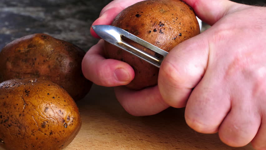 Hands peeling potatoes. A man peels potato with a potato peeler on a wooden cutting board. Peeling vegetables on the kitchen table. Ripe potato fruits are being prepared for cooking food. Close-up