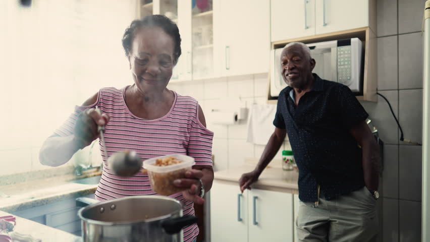 Happy Elderly African American couple in home kitchen, a black senior wife serving food into bowl standing by stove with partner in background Royalty-Free Stock Footage #1105021275