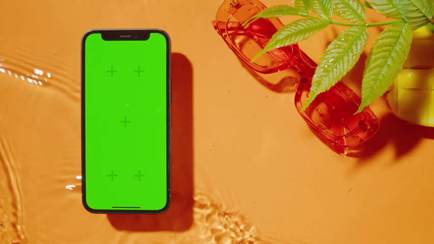 Phone with green screen chroma key on orange background. Composition of smartphone cell phone sunglasses, leaves and fruits, waves slow motion, close up, top view. Summer bright texture studio shot.