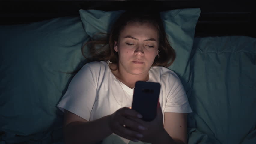 Young tired woman uses smart phone on the bed before sleeping at night. Mobile addict Royalty-Free Stock Footage #1105030139