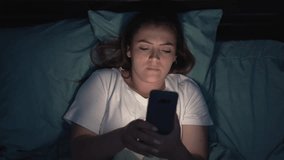 Young tired woman uses smart phone on the bed before sleeping at night. Mobile addict