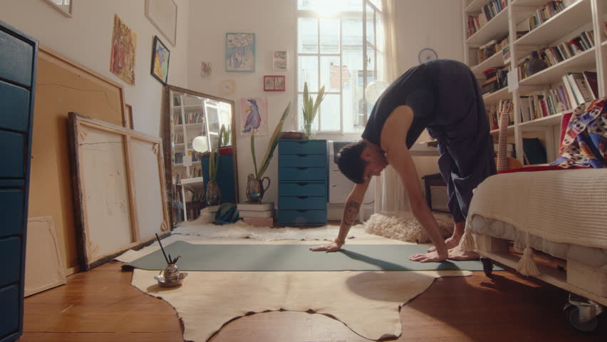 Young man in loose pants and tank top doing plank with pushups and downward dog, practicing vinyasa yoga in sunlit room with burning aroma sticks, exercising at home Royalty-Free Stock Footage #1105031733