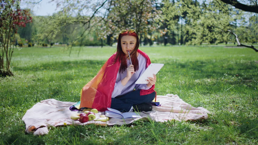 In the amazing park good looking young woman holding the lgbt flag she take some notes from her digital tablet and write on the her notebook while sitting down on the grass
