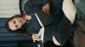 Young brunette woman reading book and listening to music at home. Vertical video. Female sitting on cozy couch at home reading book and listening to music in earphones, spending weekend at home