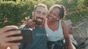 Happy smiling couple taking selfie on phone while sitting on park bench. Backlight