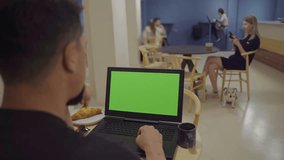 young man in cafe coworking space using laptop with green chromakey screen. Dark interior with blue wall. in the background at the table sits a blonde girl with a dog and drinks coffee. Video footage