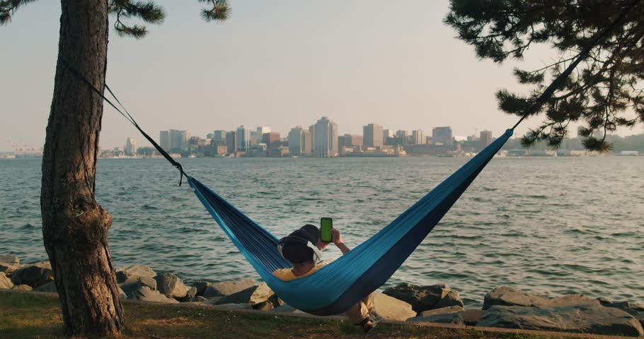 Man in a Hammock Holds a Mobile Phone Vertically with His Hand and Swipes His Finger Across the Touch Screen. Use Green Screen for Copy Space. Chroma Key Mockup on Smartphone.