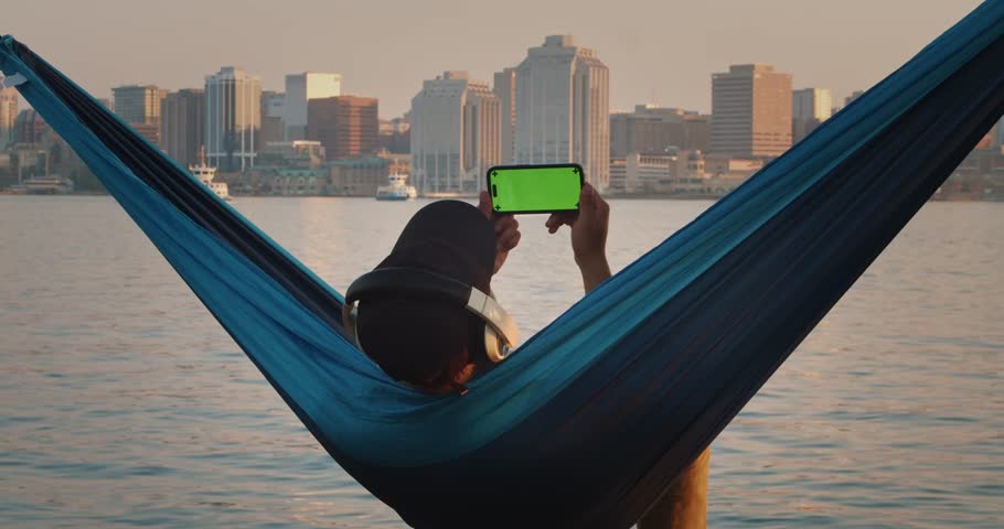 Man in a Hammock Holds a Mobile Phone Horizontally with His Hand and Swipes His Finger Across the Touch Screen. Use Green Screen for Copy Space. Chroma Key Mockup on Smartphone.