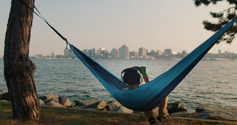 Man in a Hammock Holds a Mobile Phone Vertically with His Hand and Swipes His Finger Across the Touch Screen. Use Green Screen for Copy Space. Chroma Key Mockup on Smartphone.