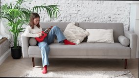 A girl is sitting in a loft-style room on a sofa, playing a red ukulele. Time for a hobby. Horizontal photo. Overall plan. Video 4K