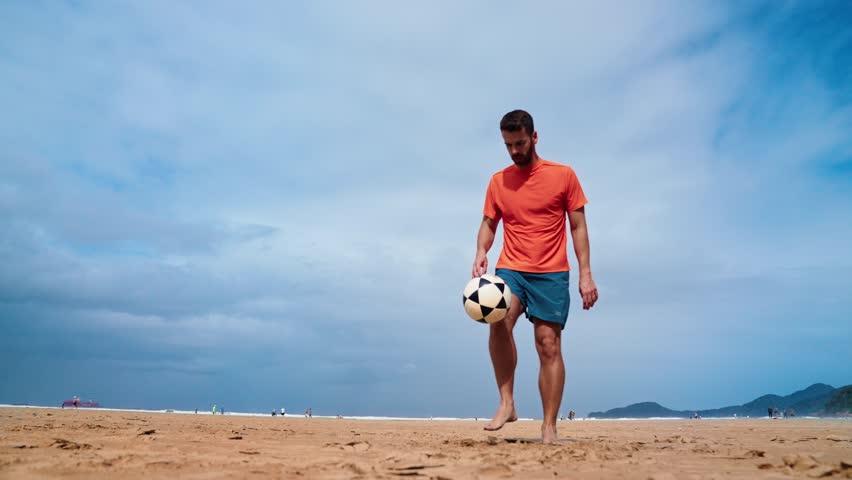 Football freestyle. Adult man practices with a soccer ball on the beach. Cinematic video of a adult man with a orange shirt playing football with the beach and the sea in the background. Royalty-Free Stock Footage #1105067411