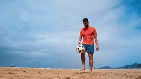 Football freestyle. Adult man practices with a soccer ball on the beach. Cinematic video of a adult man with a orange shirt playing football with the beach and the sea in the background.