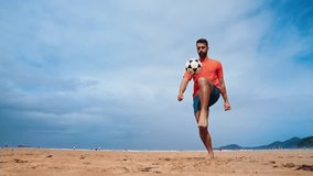 Football freestyle. Adult man practices with a soccer ball on the beach. Cinematic video of a adult man with a orange shirt playing football with the beach and the sea in the background.