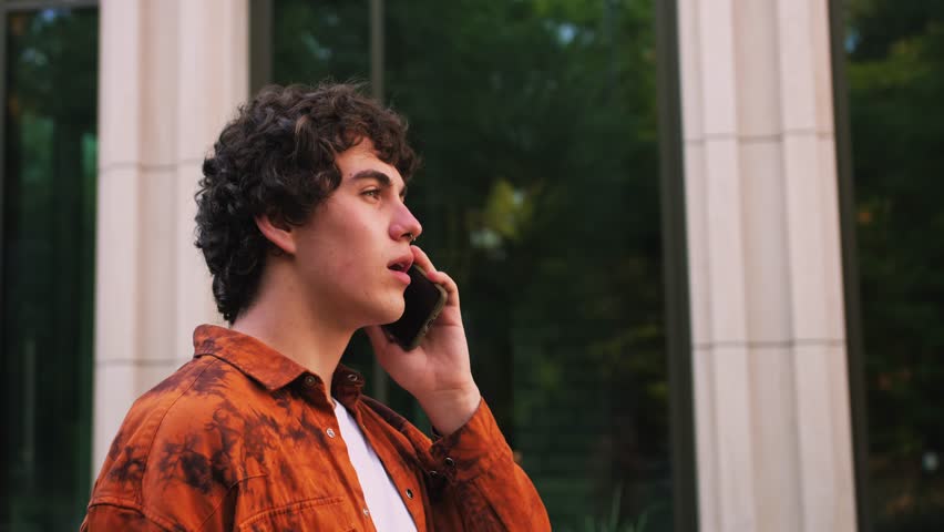 Close-up, of young man in orange shirt talking on mobile phone and walking down the street modern buildings business office background