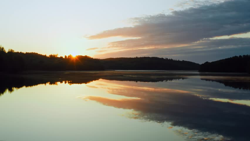 Amazing lake from above, aerial drone flies low over water surface during sunrise, beautiful island backlit with early morning sunlight. Rising sun and colored clouds in sky reflected in water mirror