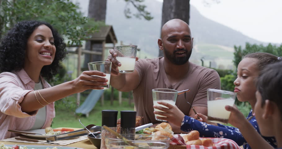 Happy biracial parents, son and daughter drinking toast at dinner table in garden, slow motion. Family, food, meal, togetherness, healthy living, lifestyle, and domestic life, unaltered.