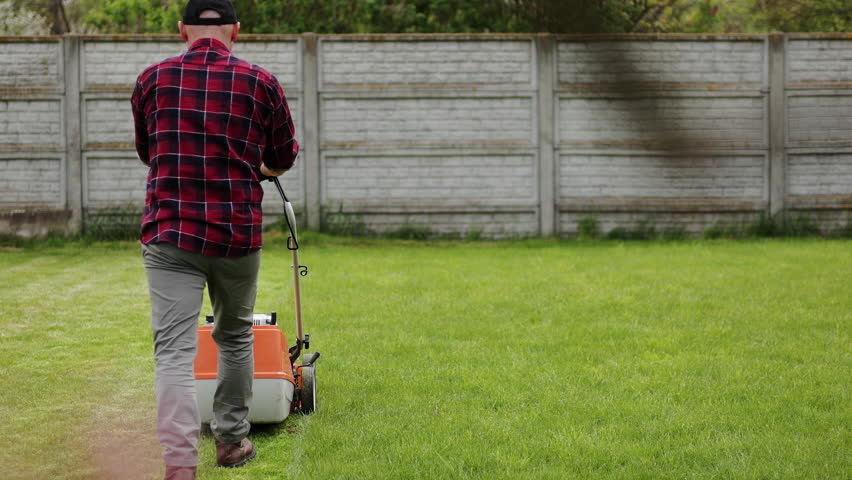 Back view of man in casual clothes mows lawn with lawn mower at backyard of his house. Husband takes care of garden on spring cloudy day. Modern gasoline garden equipment. Landscaping work. Royalty-Free Stock Footage #1105075059