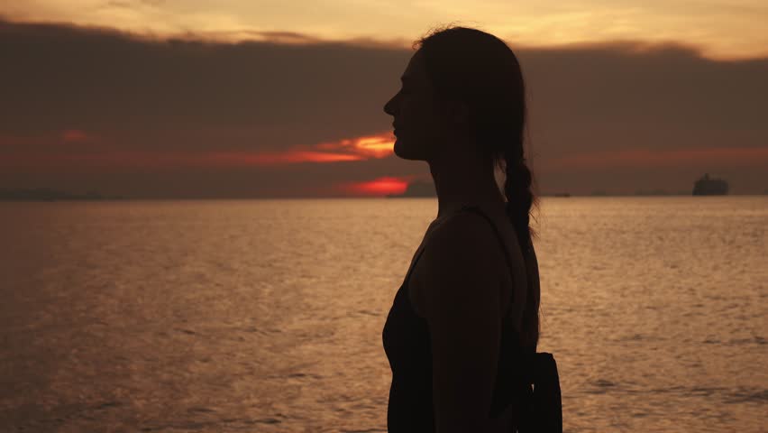 Silhouette of young female stands sideways against the backdrop of the sea with a beautiful sunset in the sky. Female smoothly raises her hands enjoying sea sunset landscape while traveling.
