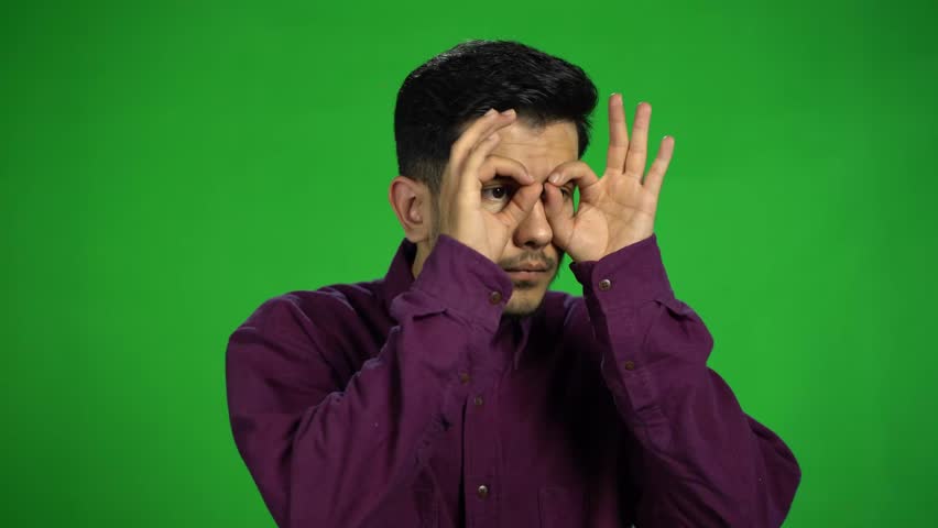 a handsome funny young Asian man making eyeglasses with hands while he is shocked and surprised , looking around , good feeling positive emotion in purple shirt over green background  Royalty-Free Stock Footage #1105088123